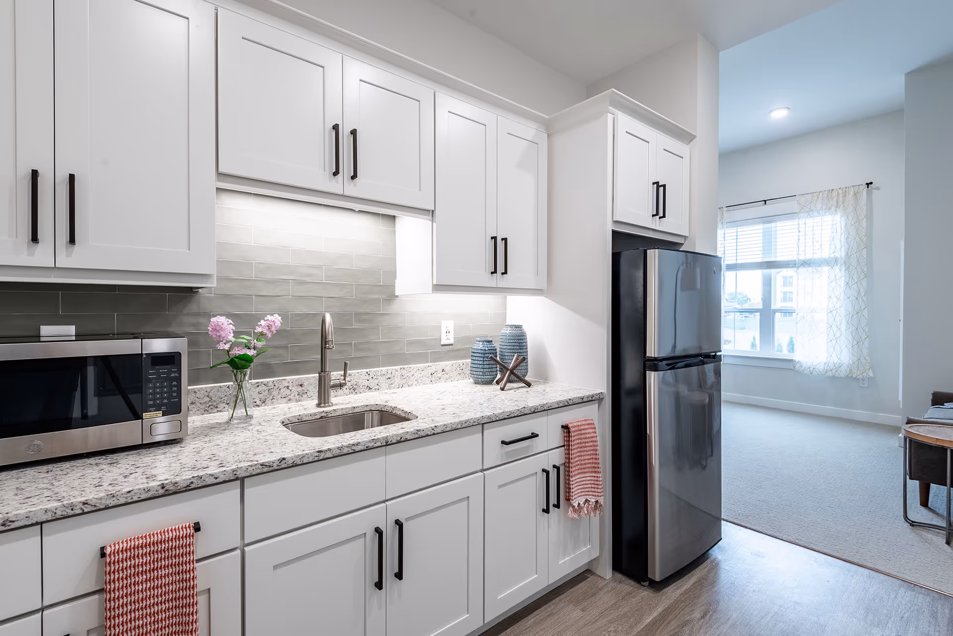 Modern white kitchen with granite countertops, a stainless steel refrigerator and microwave, sink, and decorative accents.