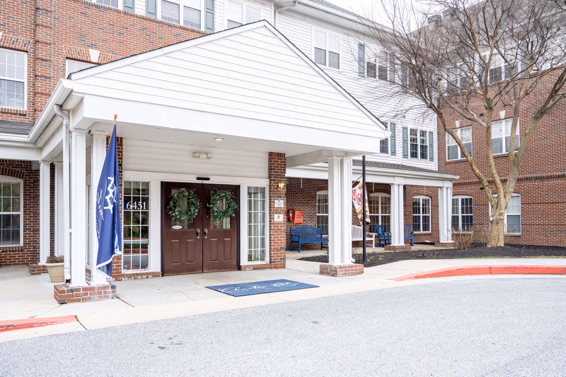 Entrance of Charter Senior Living of Towson featuring a covered drop-off area with double brown doors decorated with wreaths. The building exterior is made of red brick with white trim and multiple windows. There are two flags on either side of the entrance and blue benches under the covered porch area. Leafless trees and a paved driveway are visible in front.