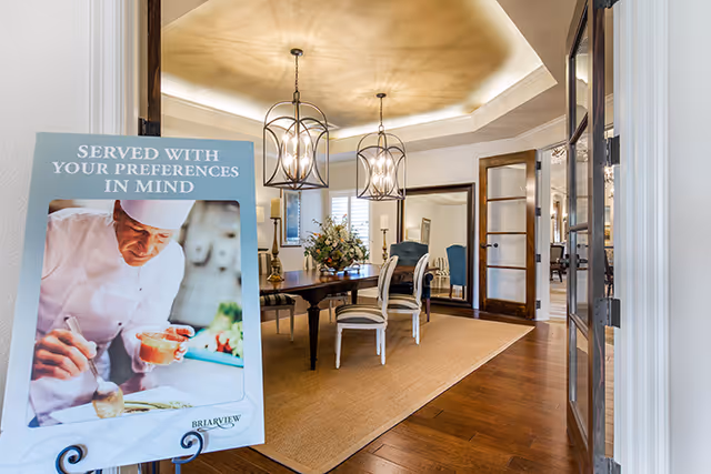 View into a dining room with a wooden table and chairs, two hanging light fixtures, a large mirror on the wall, and a floral centerpiece on the table. In the foreground, there is a sign with a chef preparing food and the text 'SERVED WITH YOUR PREFERENCES IN MIND' along with the Briarview Senior Living logo.