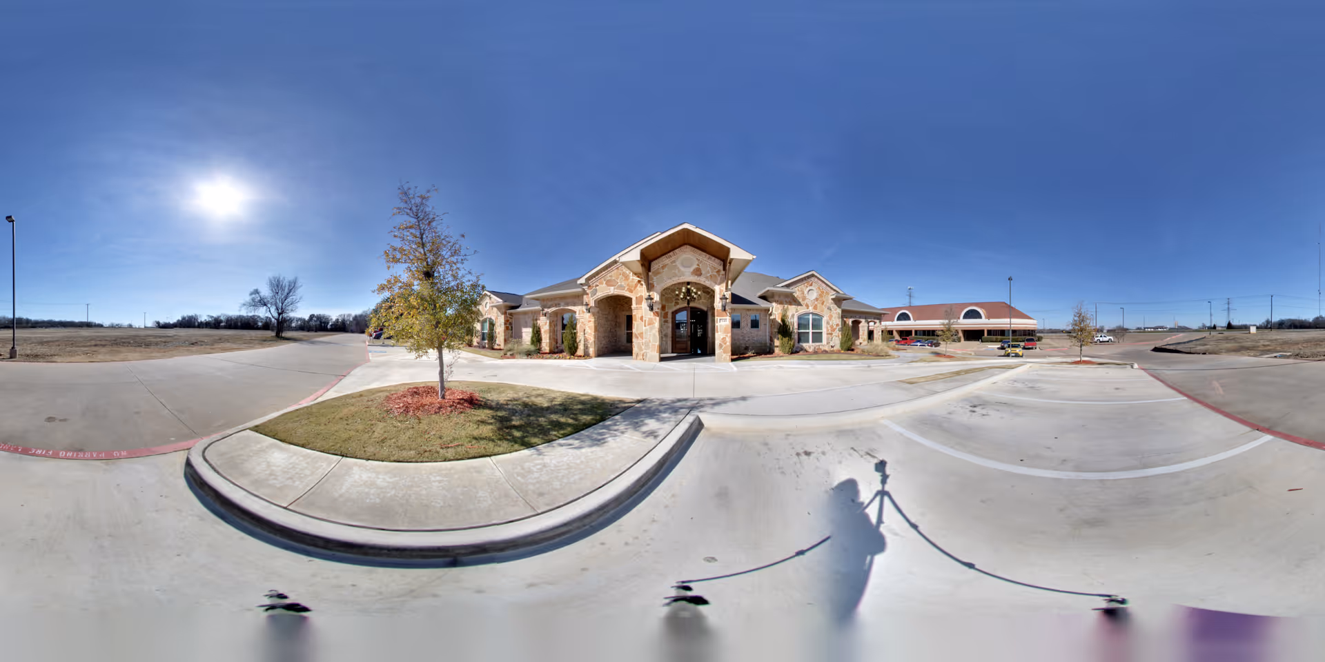 Wide panoramic view of the front exterior of Avalon Memory Care facility on a sunny day, showing a stone building with arched entrance, a small landscaped area with a tree, and a large empty parking lot under a clear blue sky.