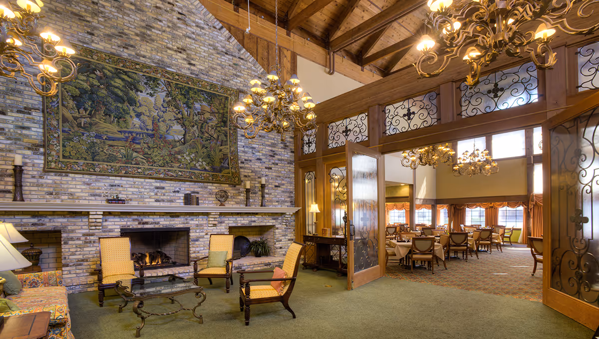 Spacious senior living common room with a stone fireplace, seating area and an adjoining dining room with chandeliers.