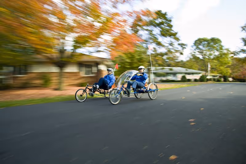Two people riding recumbent tricycles on a paved road with trees showing autumn colors and residential buildings in the background.