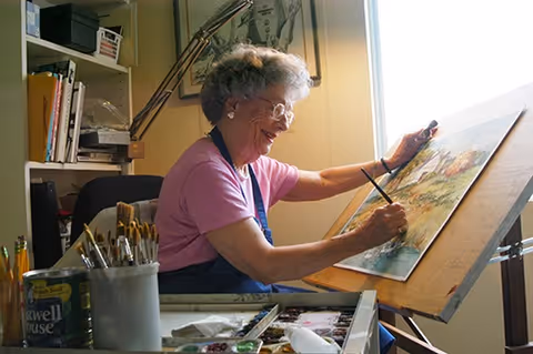 An elderly woman with gray hair and glasses is smiling while painting on a large canvas set on an easel in a well-lit room. She is wearing a pink shirt and a dark apron. The room has shelves with art supplies and books, and there are paintbrushes and painting materials on the table in front of her.