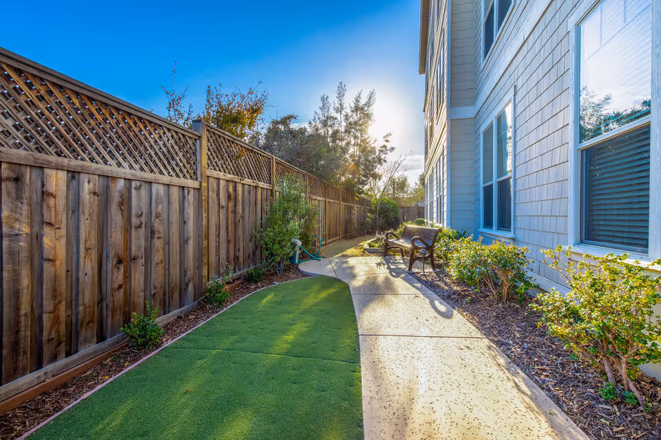Outdoor pathway alongside a building with a wooden fence on one side and bushes on the other. There is a bench along the pathway and the sun is shining brightly in the background.