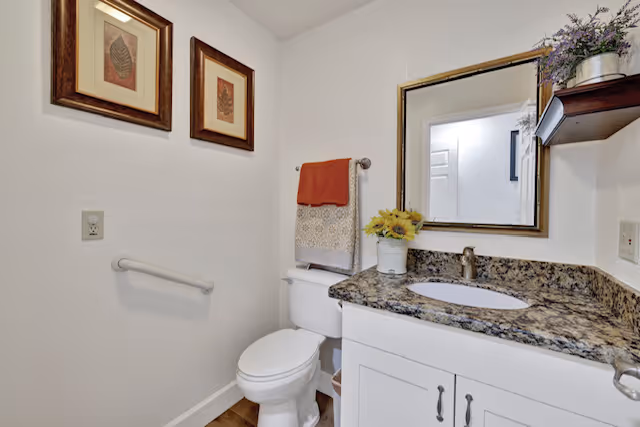 Small bathroom with a toilet, white vanity with granite countertop and sink, mirror, towel, and framed wall art.