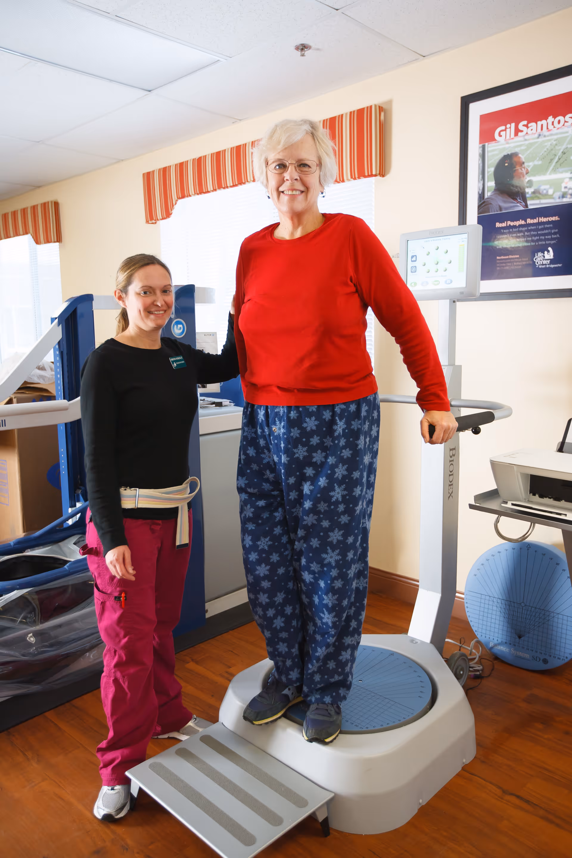 An elderly woman wearing a red sweater and blue patterned pants stands on a Biodex balance testing machine while a female healthcare worker in a black top and pink pants stands beside her, providing support. The setting appears to be a rehabilitation or physical therapy room with medical equipment and a framed poster on the wall.