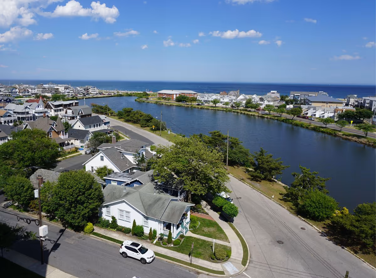 Aerial view of a coastal neighborhood with houses, a road, a waterway, and the ocean in the background under a blue sky with scattered clouds.