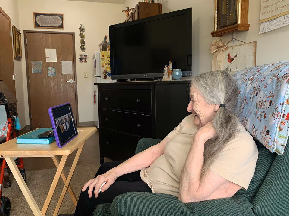 An elderly woman with gray hair tied in a ponytail is sitting on a green armchair in a cozy room. She is smiling and looking at a tablet on a small wooden table in front of her. The room has a black dresser with a TV on top, a door with decorations, and various wall hangings including a calendar and a sign that says 'Duckworth'.