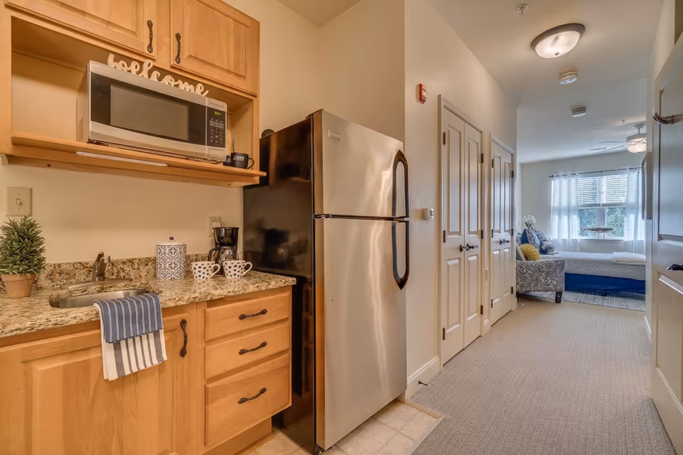 Interior view of a senior living facility unit showing a small kitchen area with wooden cabinets, a granite countertop with a sink, a microwave, coffee maker, and a stainless steel refrigerator. The hallway leads to a bedroom with a bed, a window with white curtains, and a chair with cushions.