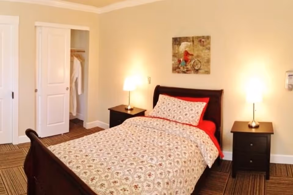 A neatly furnished bedroom with a single bed, patterned bedding, two nightstands with lamps, an open closet, and a painting on the wall.