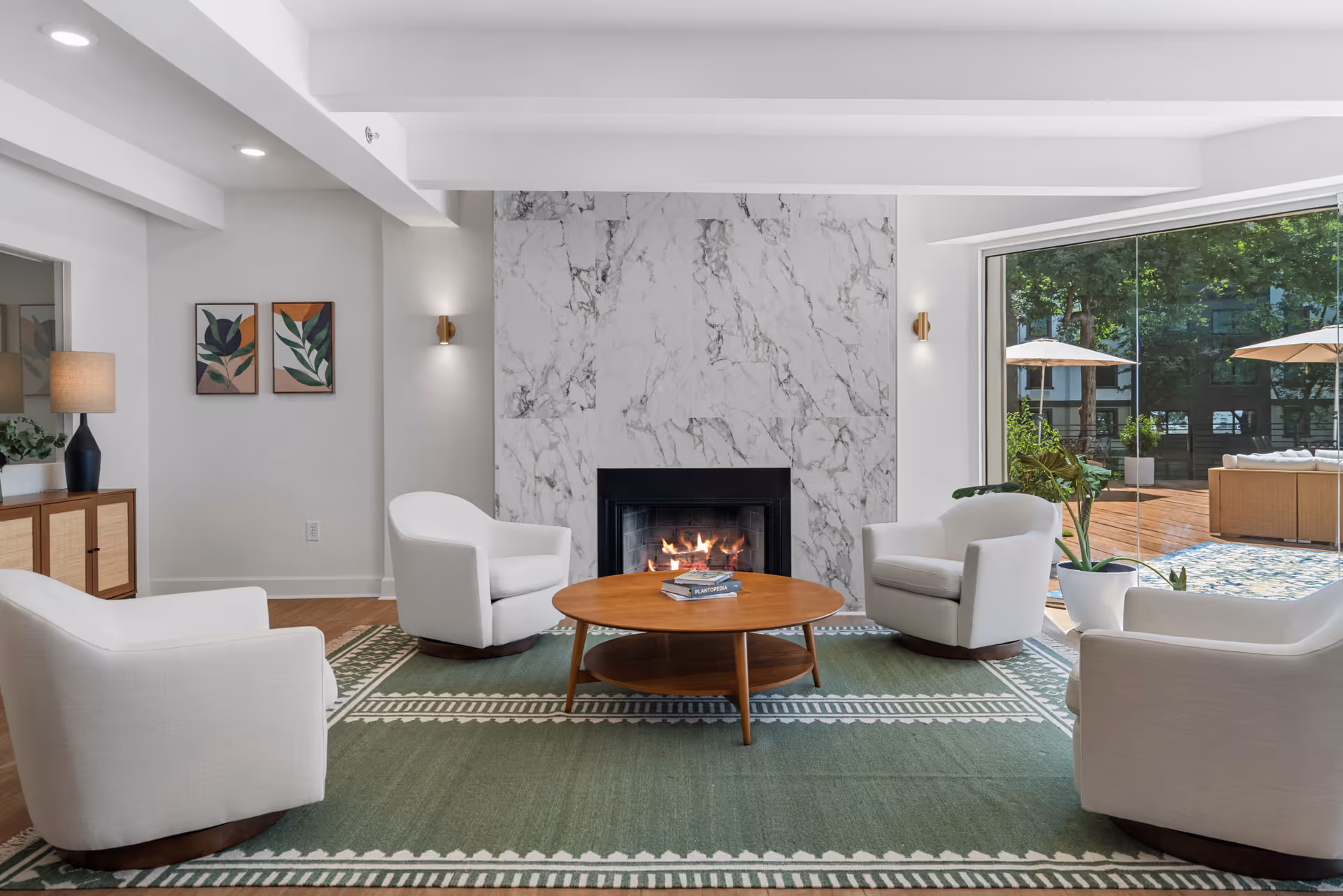 A modern living room with four white armchairs arranged around a round wooden coffee table on a green patterned rug. A fireplace with a marble surround is centered on the back wall, and a large glass sliding door on the right opens to an outdoor patio with umbrellas and seating. The room also features a wooden sideboard with a lamp and two framed botanical prints on the wall.