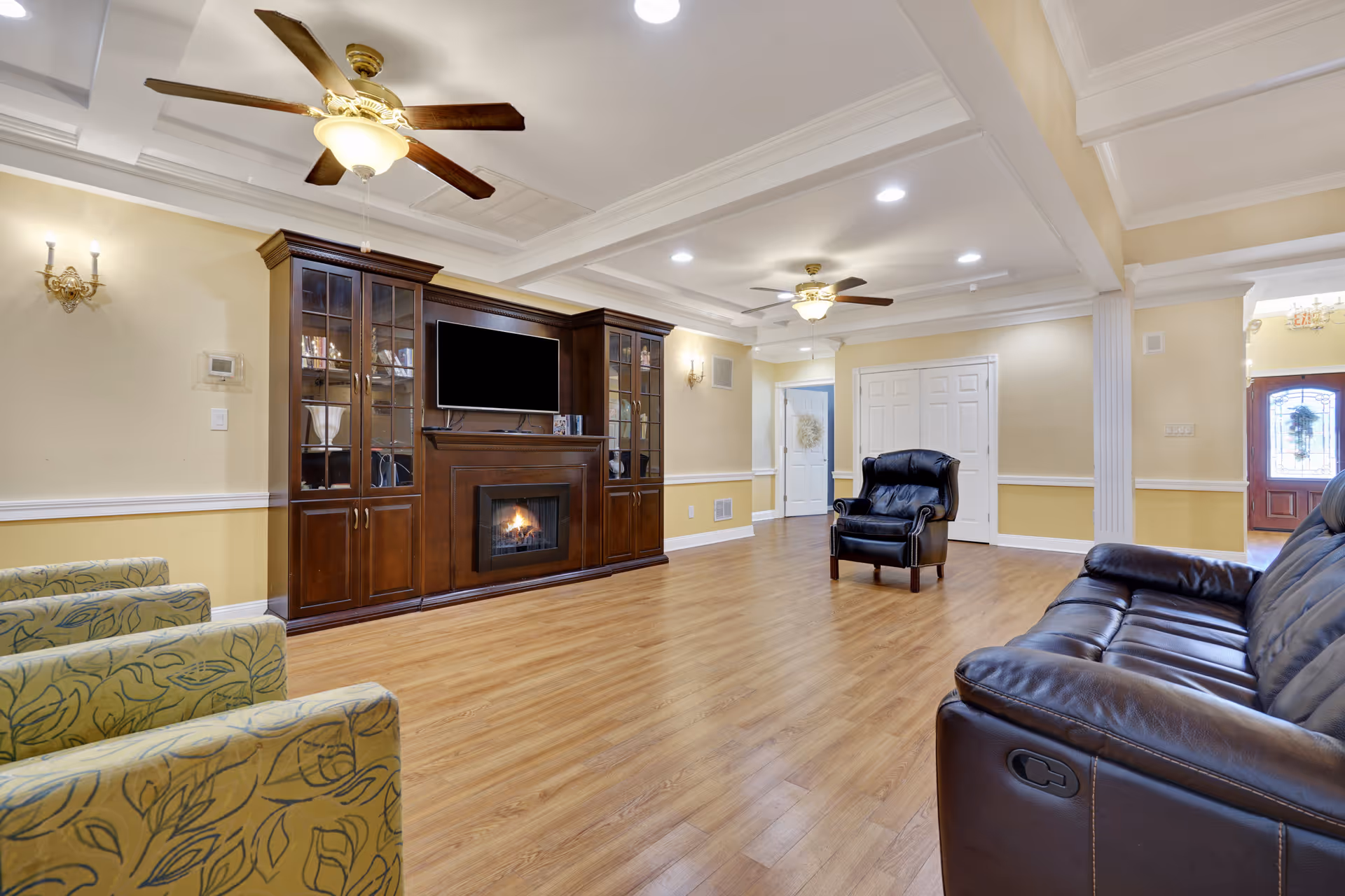 A spacious living room with wooden flooring, beige walls with white trim, and a coffered ceiling with recessed lighting and ceiling fans. The room features a dark wooden entertainment center with a fireplace and a mounted flat-screen TV. There are several seating options including a black leather sofa, a black leather armchair, and two patterned armchairs. A doorway and an entrance with a glass-paneled door are visible in the background.