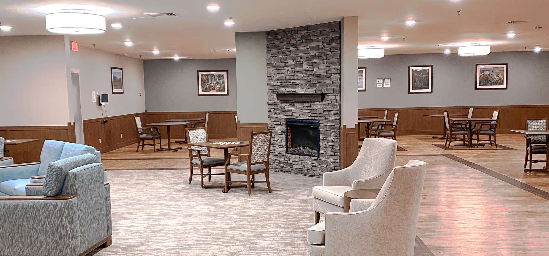 A spacious common area in a senior living facility featuring a stone fireplace in the center, surrounded by multiple seating arrangements including cushioned armchairs and wooden tables with chairs. The walls are decorated with framed artwork, and the room is well-lit with ceiling lights.