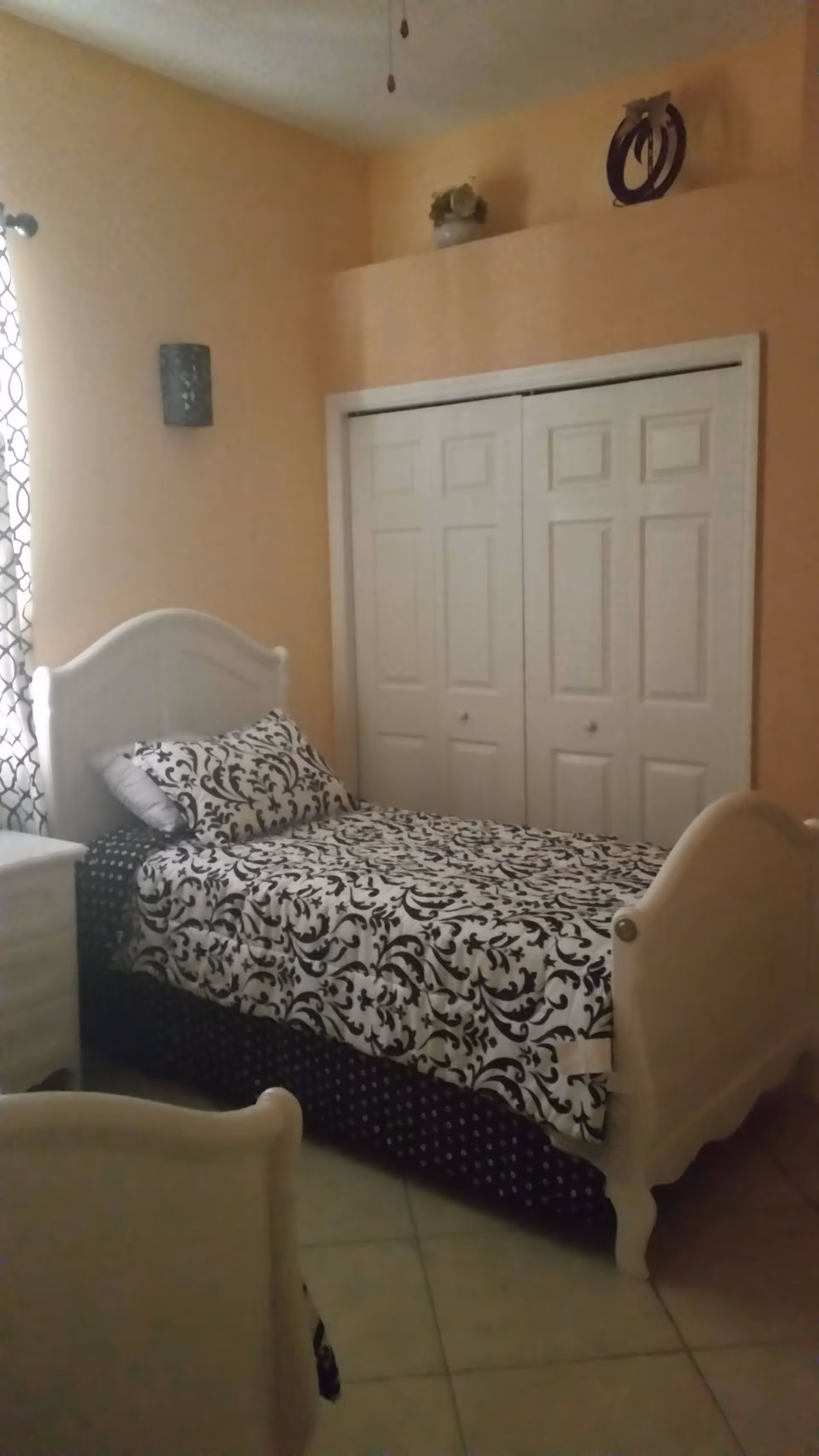 A small bedroom with a single white bed featuring a black and white patterned bedspread and pillow. The room has beige walls, a white nightstand, a window with patterned curtains, and a double-door closet. Decorative items are placed on a ledge above the closet.