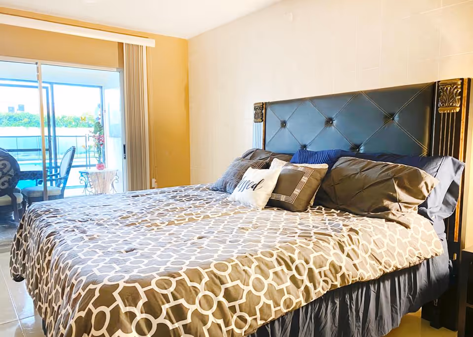 A bedroom with a large bed featuring a patterned bedspread and multiple pillows, including one with the word 'Blessed' on it. The room has a sliding glass door leading to an outdoor patio area with chairs and a table. The walls are light-colored, and the headboard of the bed is dark with decorative gold accents.