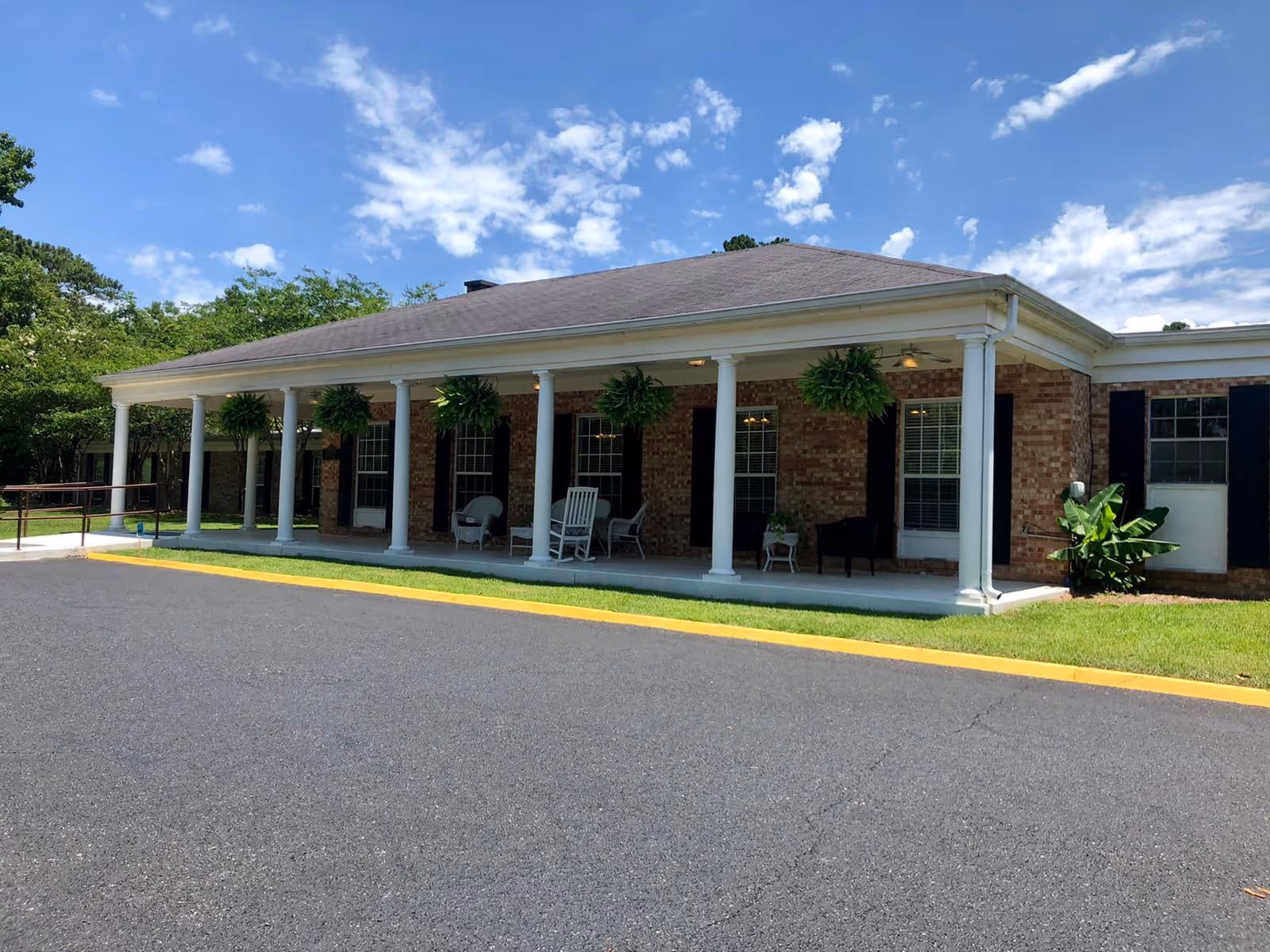 Exterior view of a single-story brick building with a covered porch supported by white columns. The porch has several hanging plants and outdoor chairs, with a clear blue sky and scattered clouds above.