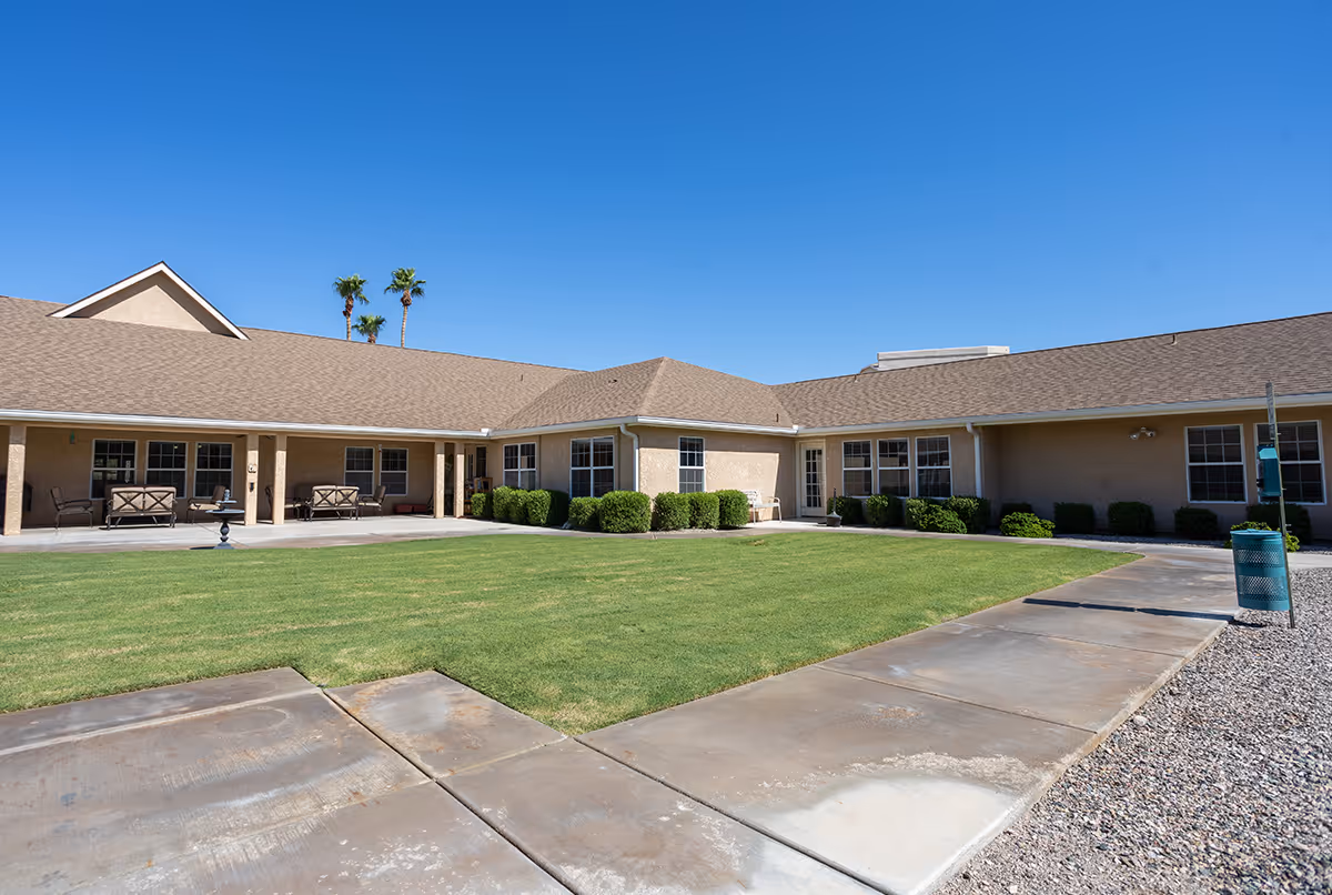 Outdoor courtyard area of Avista Senior Living Lake Havasu with a well-maintained green lawn, concrete walkways, beige building with multiple windows, patio seating, and clear blue sky.