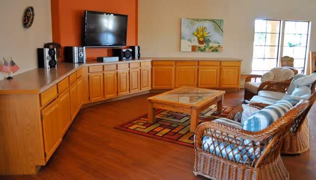 A cozy living room area with wicker furniture including a sofa and chairs with cushions, a wooden coffee table on a colorful patterned rug, a wall-mounted flat screen TV above wooden cabinets, and a painting on the wall. Large windows allow natural light to fill the room.
