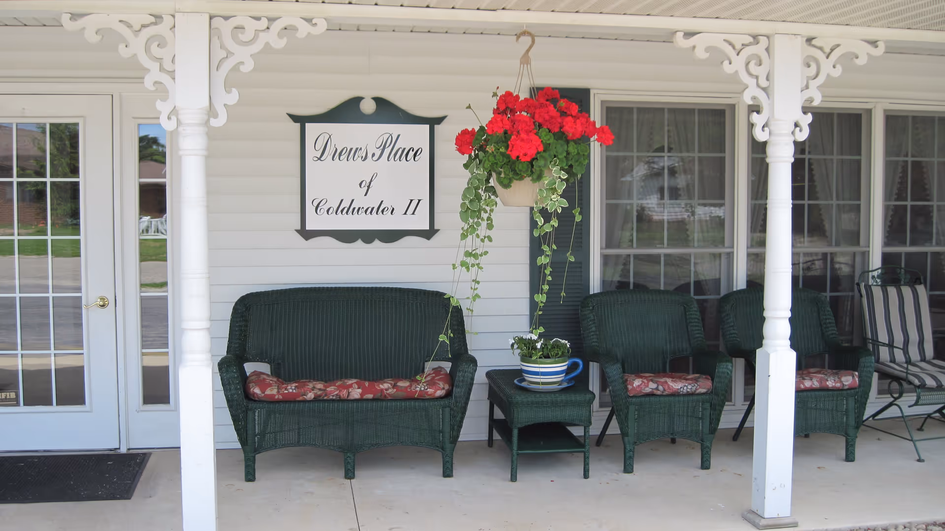 Front porch area of Drews Place of Coldwater II featuring green wicker seating with floral cushions, a small table with a potted plant, a hanging basket with red flowers, white decorative pillars, and a sign displaying the facility name.