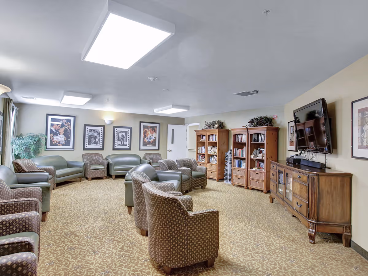 A spacious senior living lounge with multiple armchairs and sofas, bookshelves, framed art, and a wall-mounted TV.