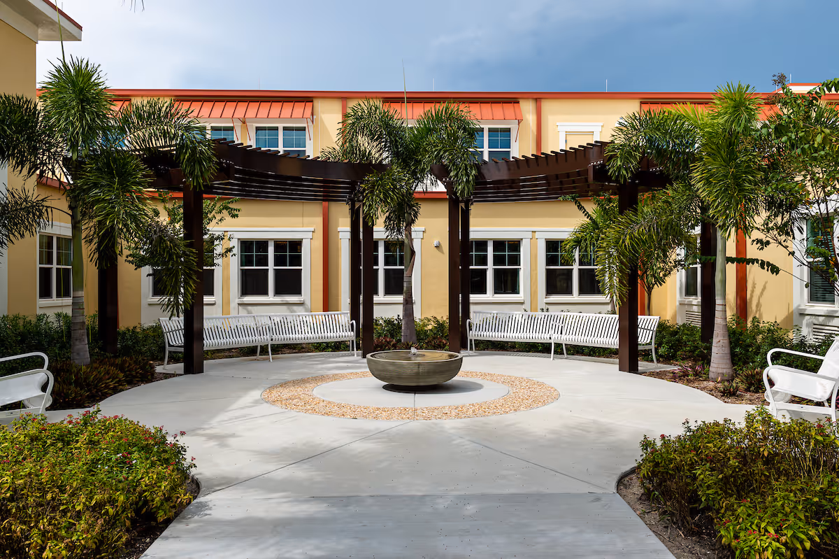 Courtyard with a central fountain, white benches, a wooden pergola and palm trees in front of a yellow building.