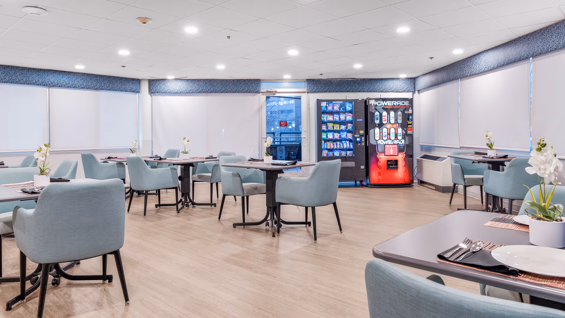 A bright dining room with multiple tables and light blue upholstered chairs arranged neatly. Each table has a small white potted plant and place settings with plates, utensils, and napkins. The room has large windows covered with white blinds and a light wood floor. In the background, there are two vending machines, one for snacks and one for drinks. The ceiling has recessed lighting and a blue patterned valance runs along the top of the windows.