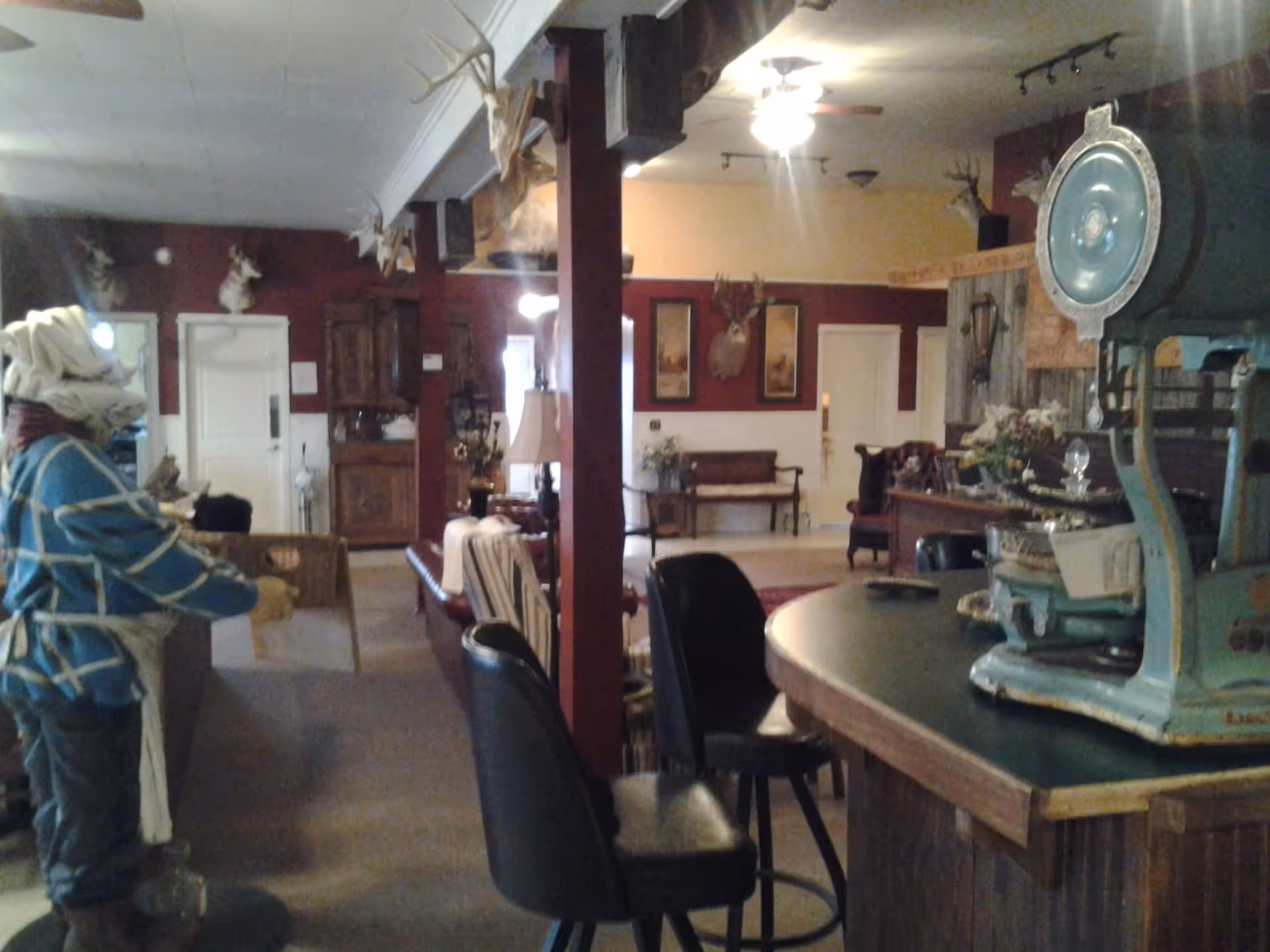 Interior view of a rustic common area with wooden furniture, mounted deer heads on the walls, a vintage scale on a counter, and a mannequin dressed in western attire. The room has a warm, cozy atmosphere with a mix of dark red and beige walls and ceiling fans.