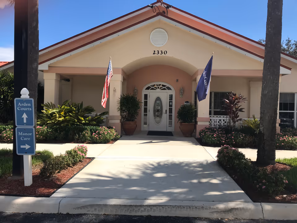 Front entrance of a single-story building with the address number 2330 above the door. The entrance has a white door with decorative glass panels and is flanked by two large potted plants. Two flags, one American and one with the text 'Arden Courts,' are displayed on either side of the entrance. There is a signpost on the left side of the walkway indicating directions to Arden Courts and Manor Care. The building is surrounded by landscaped bushes and flowers, with palm trees visible on either side.