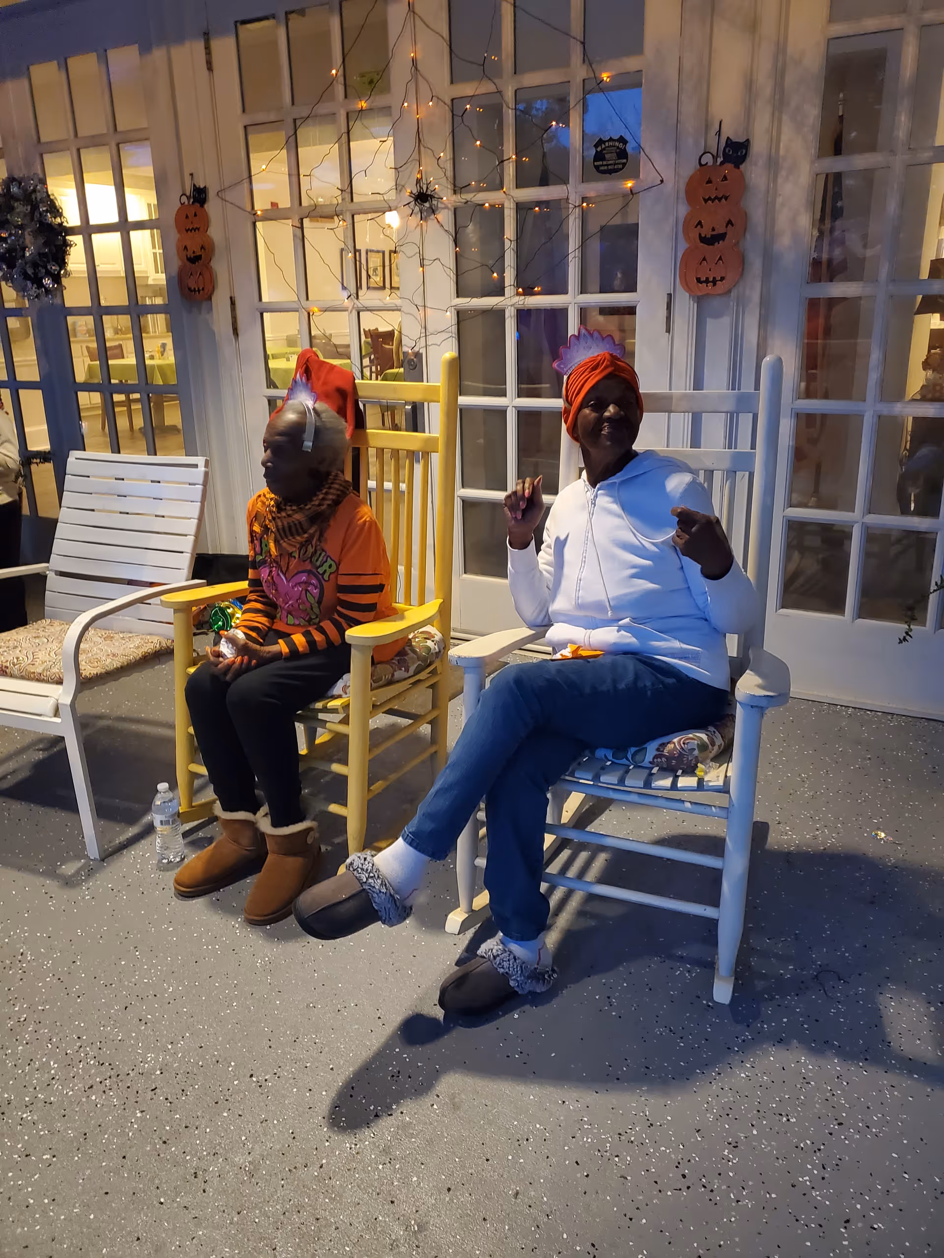 Two elderly women sitting on rocking chairs outside a building decorated with Halloween-themed items including pumpkin cutouts and a spider web with lights. One woman is wearing a red hat and an orange striped shirt, while the other is wearing a white jacket and a red head wrap. There are glass doors behind them with visible indoor lighting.