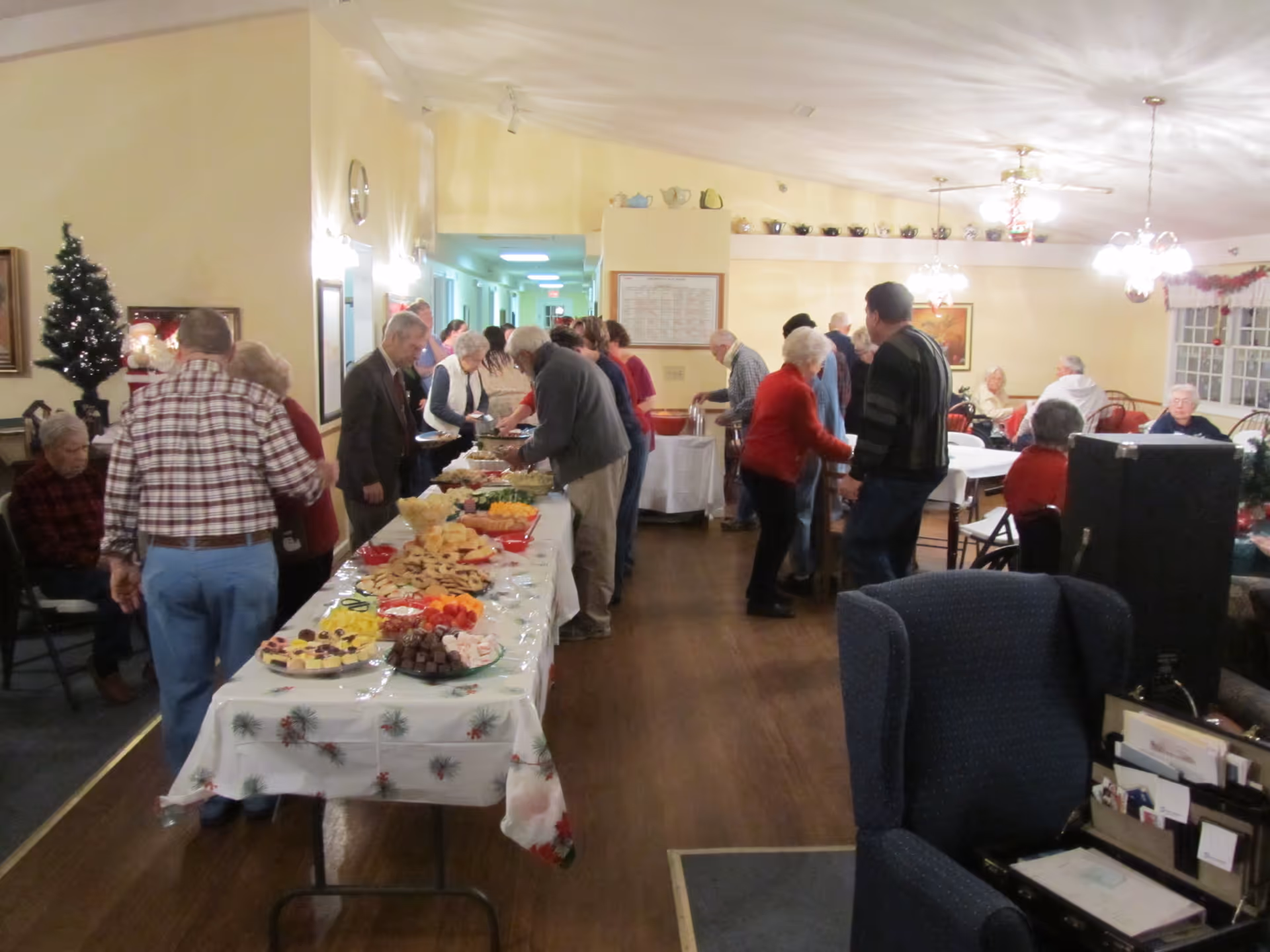 A group of elderly people gathered in a decorated common room with a long table covered with a festive tablecloth and various snacks and desserts. Some people are standing in line serving themselves food, while others are seated at tables in the background. The room has warm lighting, ceiling fans, and holiday decorations including a small Christmas tree.