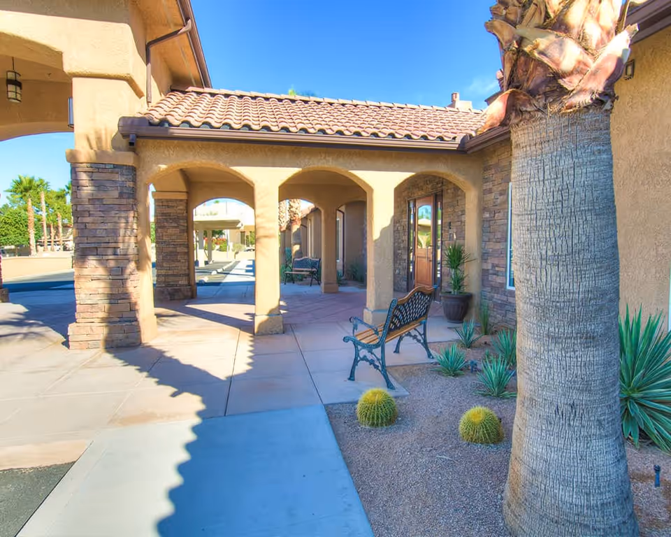 Outdoor covered walkway at Lake View Terrace Memory Care Residence with stone pillars, benches, desert plants including cacti and a palm tree, and a clear blue sky.