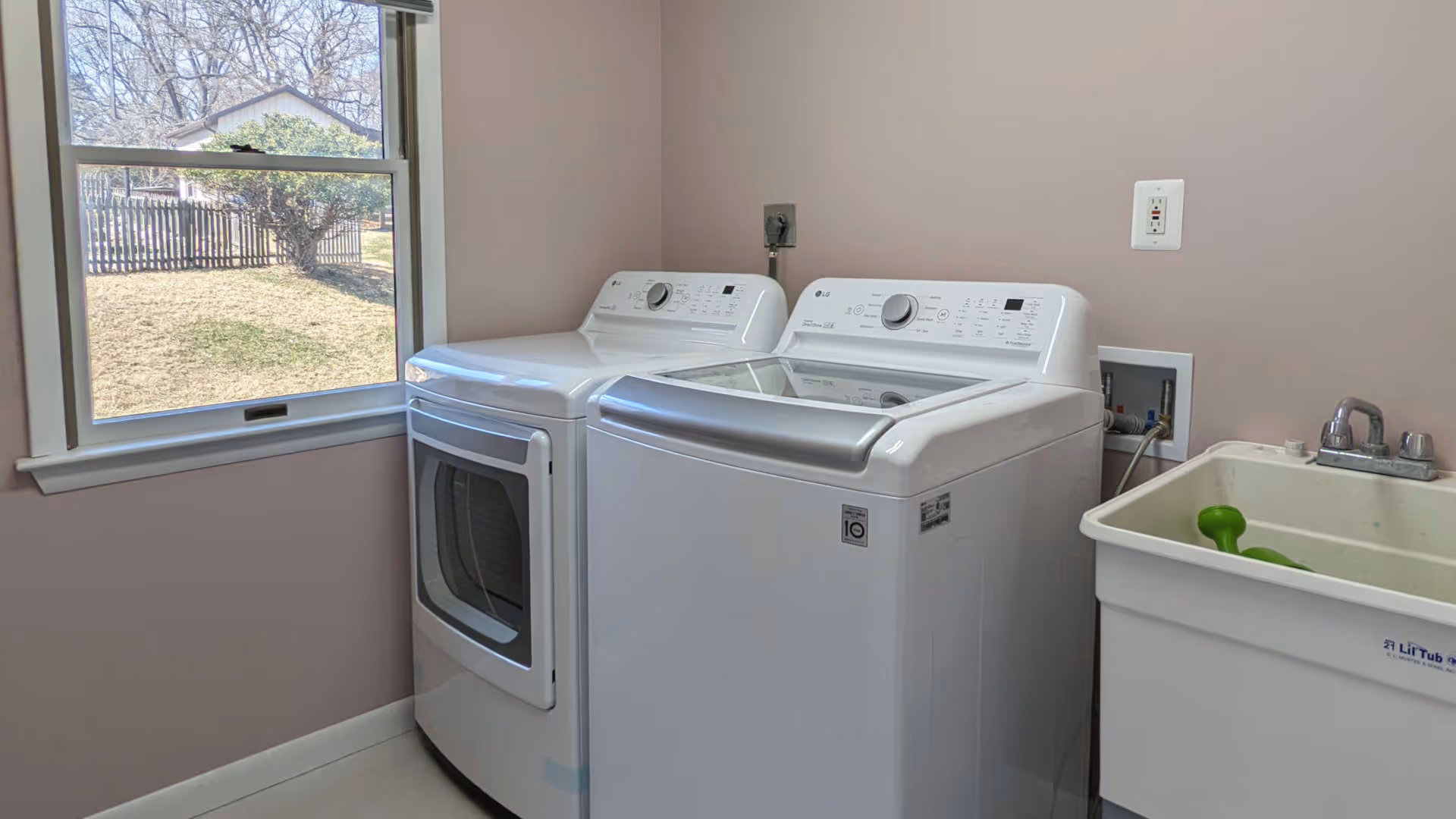 Laundry room with a white washing machine and dryer side by side next to a utility sink with a green object inside. A window shows an outdoor view with grass, a tree, and a fence.