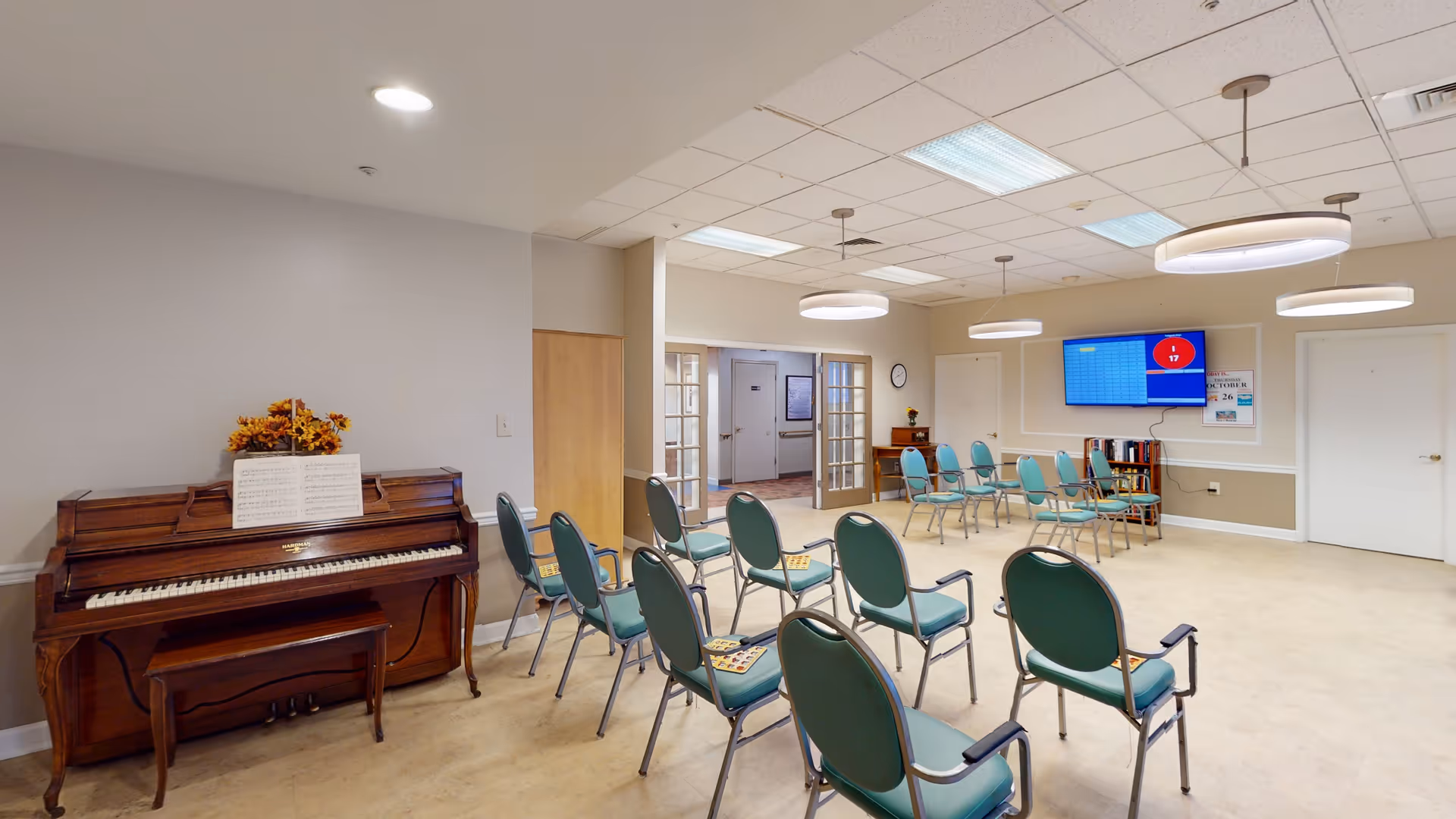 A spacious room with several green chairs arranged in rows facing a wall-mounted flat screen TV. There is a wooden piano with sheet music and a flower arrangement on top against one wall. The room has a beige carpet, white walls with a beige lower section, and a ceiling with recessed lighting and hanging circular light fixtures. There are double glass doors leading to another area and a small bookshelf beneath the TV.