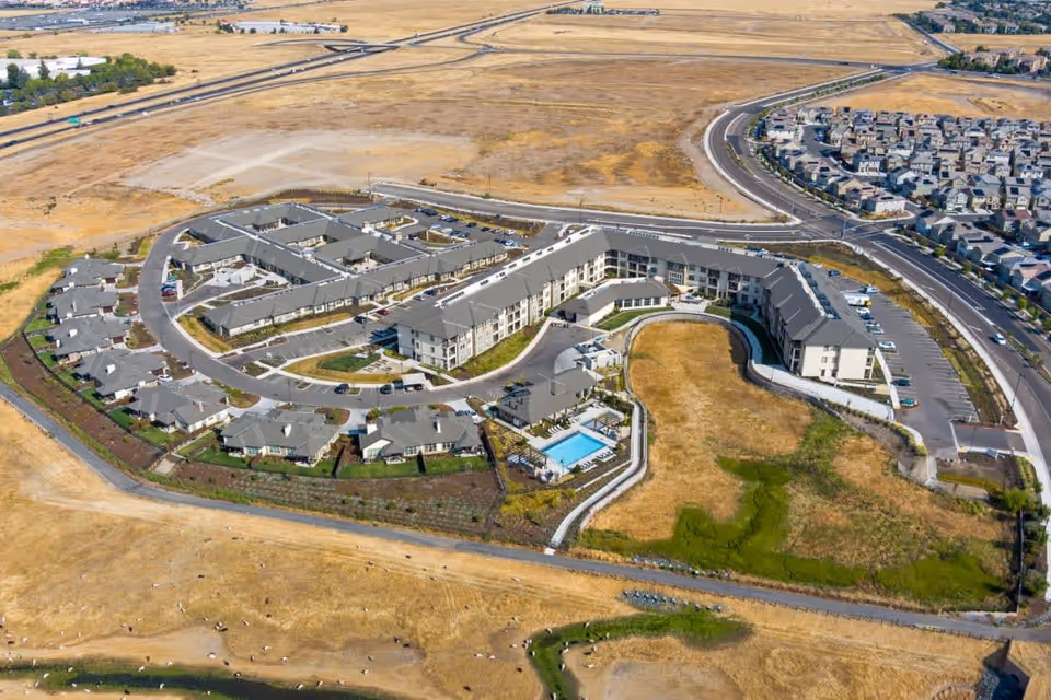 Aerial view of Ansel Park Independent Living facility showing multiple buildings arranged in a curved layout with a swimming pool, parking lots, and surrounding dry grassy land. Nearby residential neighborhood is visible in the background.