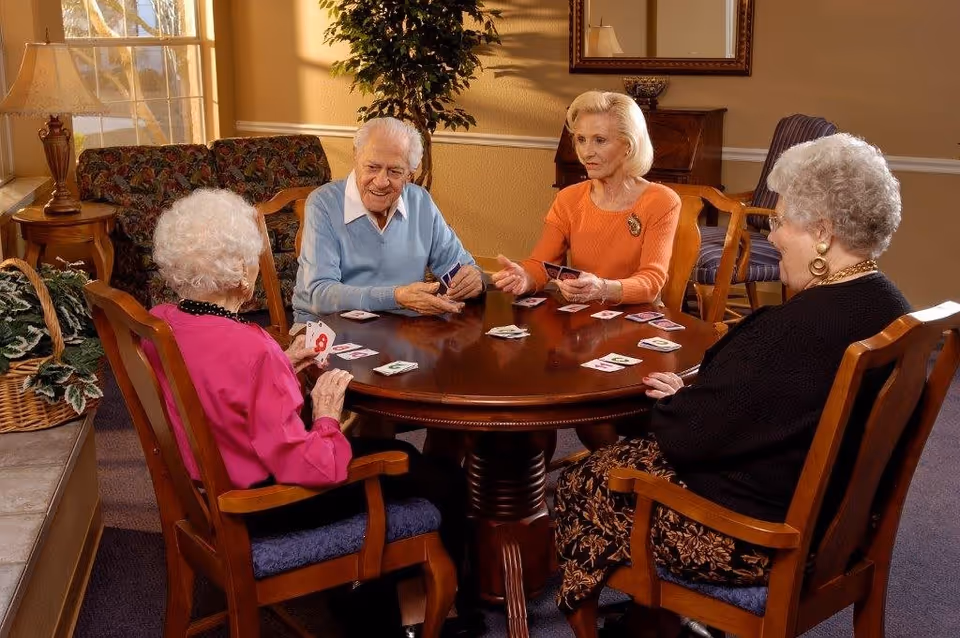 Four elderly individuals sitting around a wooden round table playing a card game in a warmly lit room with a window, lamp, plant, and framed mirror on the wall.