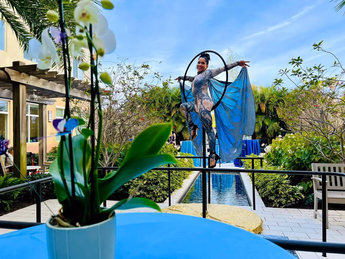A performer in a butterfly costume poses on an aerial hoop above a reflecting pool in an outdoor courtyard with potted orchids in the foreground.