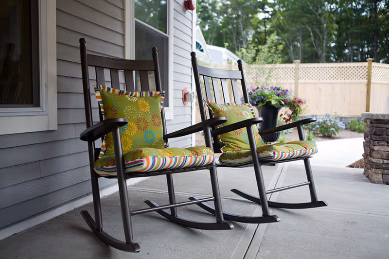 Two dark wooden rocking chairs with colorful striped and floral cushions placed on a covered concrete patio next to a gray building wall, with a wooden fence and green trees in the background.