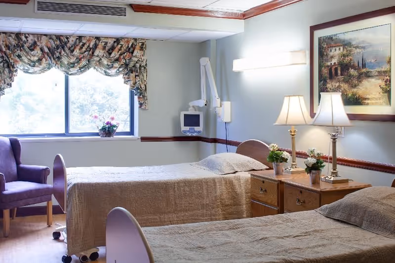 A bright shared bedroom with two twin beds, bedside tables with lamps and potted flowers, a window with a floral valance and an armchair.