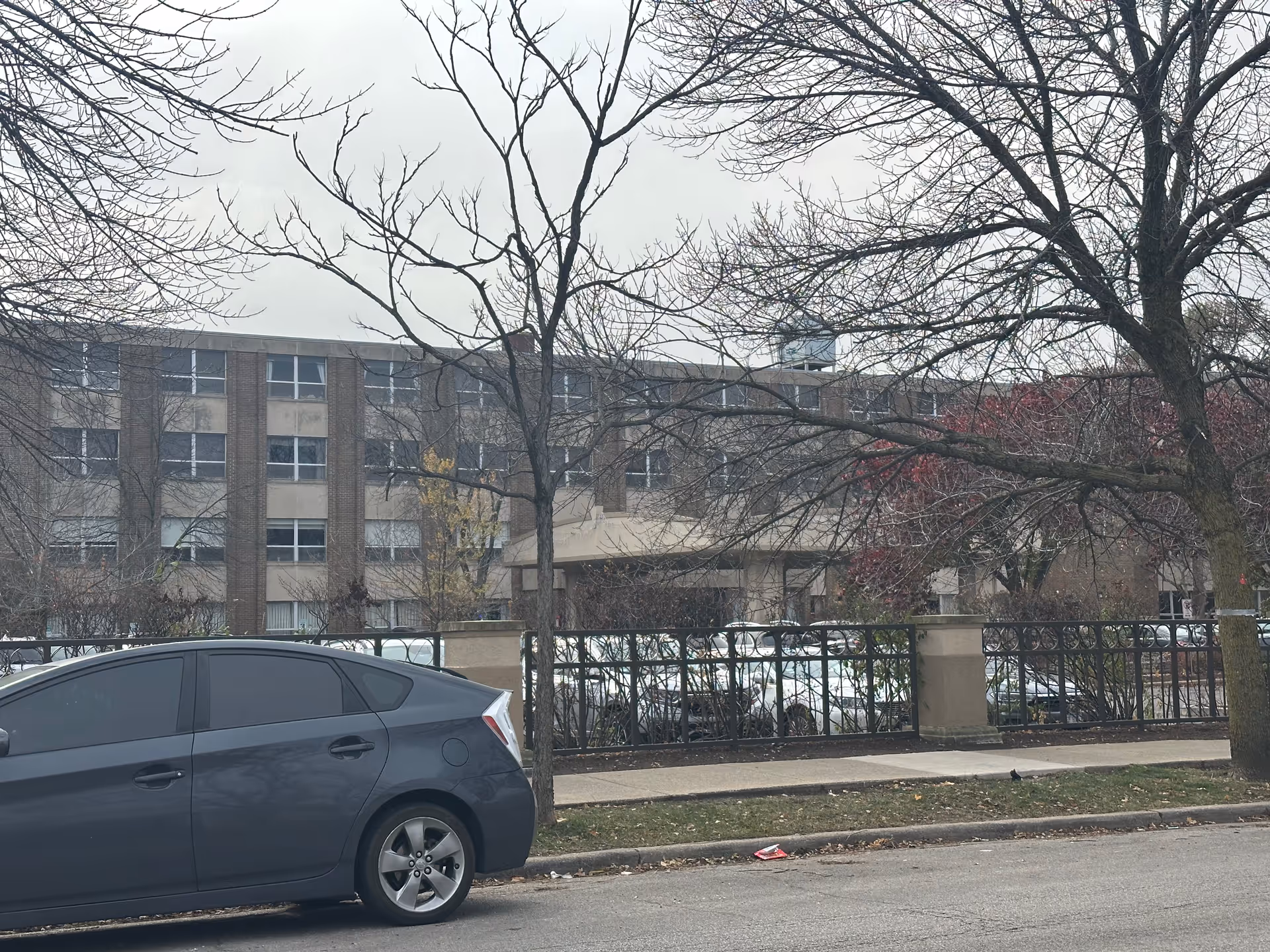 Exterior view of a multi-story brick building behind a black metal fence with leafless trees in front. A dark gray car is parked on the street in the foreground under an overcast sky.