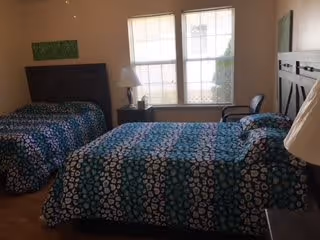 A bedroom with two twin beds featuring dark wooden headboards and blue patterned bedspreads. Between the beds is a nightstand with a white lamp and a window behind it letting in natural light. There is a chair next to the window and a piece of wall art above one of the beds.