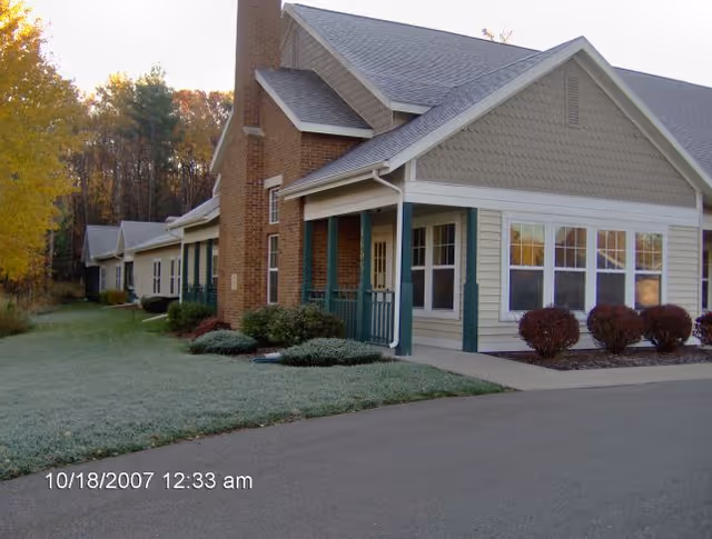 Exterior view of a single-story residential building with beige siding, green trim, and a brick chimney. The building is surrounded by a lawn with bushes and trees showing autumn colors. A paved driveway runs alongside the building.