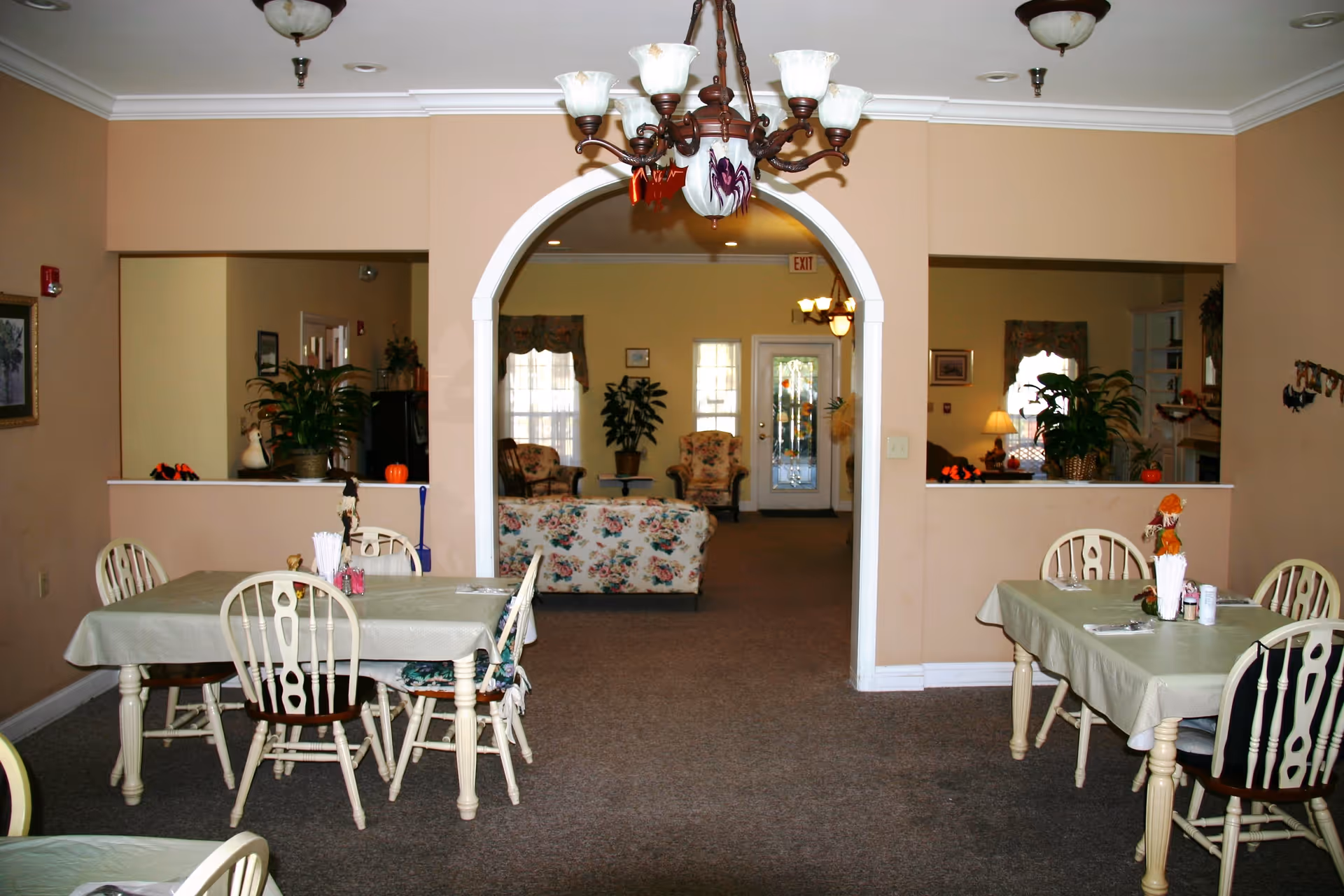 Interior view of a senior living facility dining area with several tables covered with light green tablecloths and white wooden chairs. The room has beige walls and carpeted floors. Through an arched doorway, a sitting area with floral upholstered chairs and a sofa is visible, along with plants and a glass door at the back. Ceiling light fixtures and a chandelier hang from the ceiling.