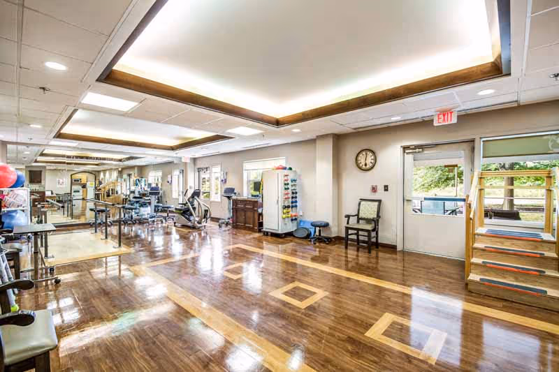 A spacious rehabilitation and therapy room with polished wooden floors, exercise equipment, therapy balls, and parallel bars. There is a clock on the wall, a door leading outside, and a set of stairs with handrails. The room is well-lit with ceiling lights and has large mirrors along one wall.