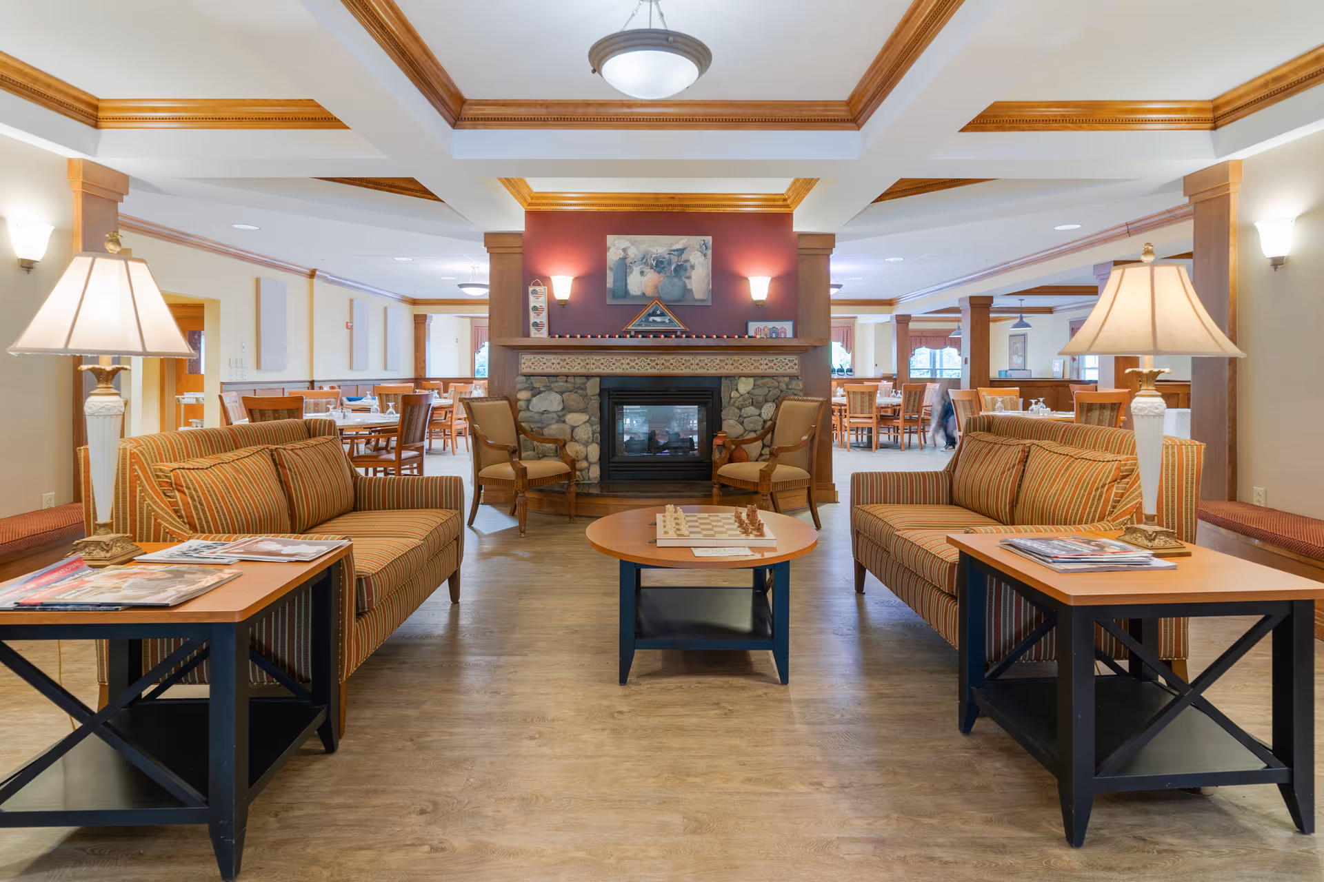 A cozy common area in a senior living facility featuring two striped sofas facing each other with wooden side tables and lamps. In the center is a round coffee table with a chessboard. Behind the seating area is a stone fireplace with a red accent wall and decorative artwork above it. The background shows a dining area with wooden tables and chairs, illuminated by ceiling lights.