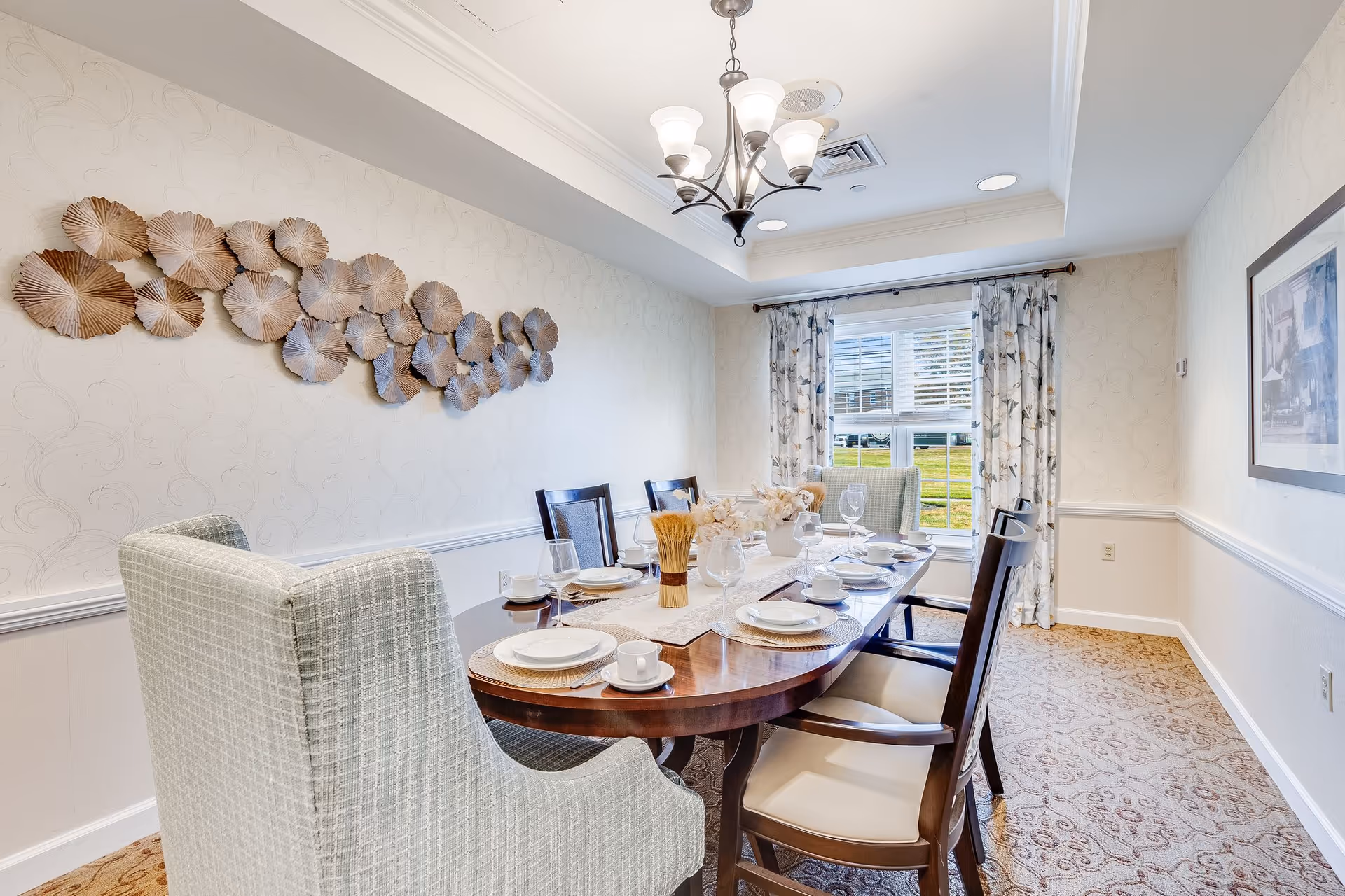 A well-lit dining room with a wooden dining table set for six, including plates, cups, and wine glasses. The room features patterned carpet, light-colored wallpaper, a decorative metal wall art piece, a chandelier, and a window with floral curtains letting in natural light.