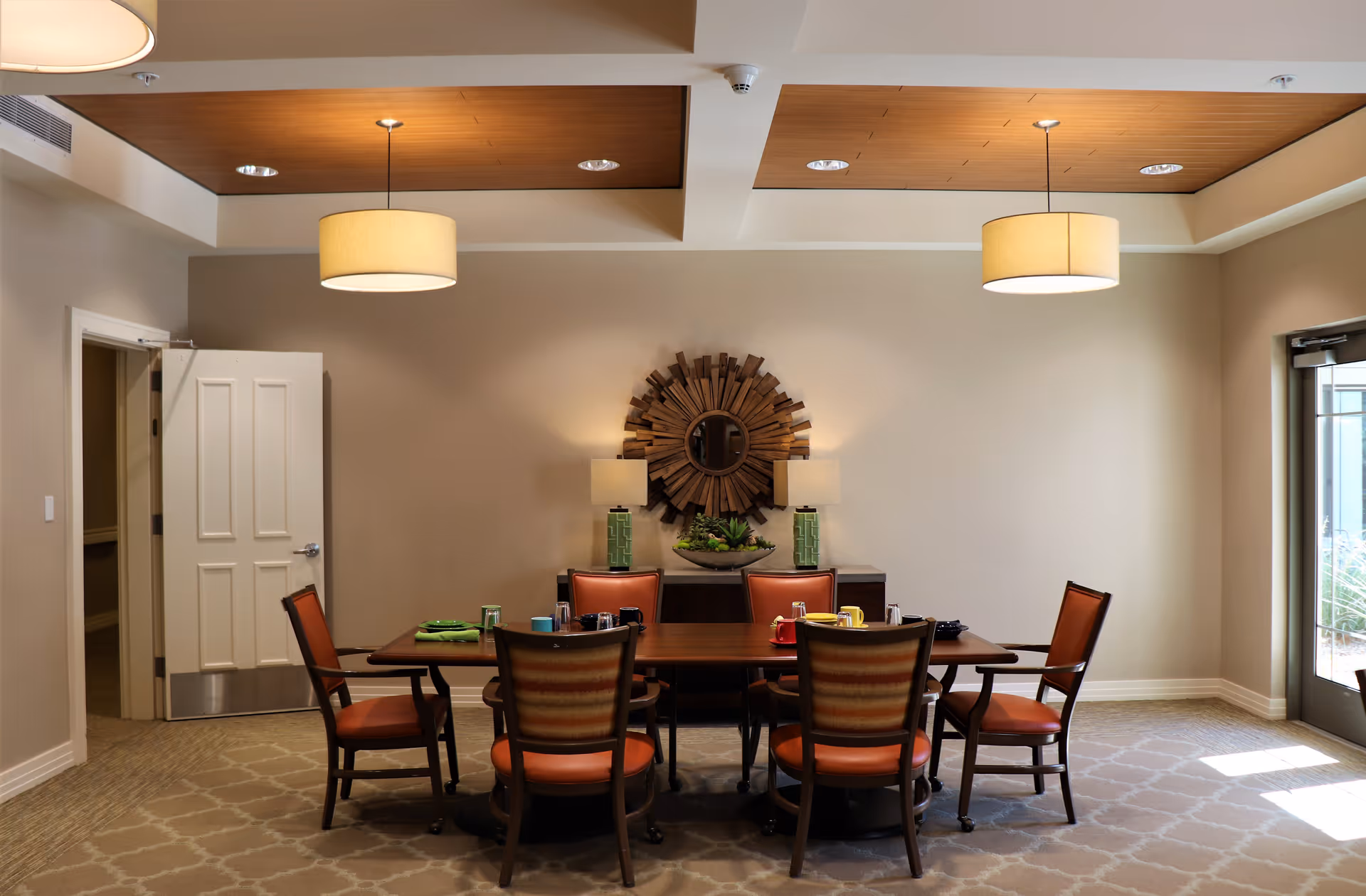 A dining room with a rectangular wooden table surrounded by six chairs with orange cushions. The room has beige walls, a patterned carpet, two hanging ceiling lamps, and a decorative wooden sunburst mirror on the wall above a sideboard with two table lamps and a plant. There is a door on the left and a glass door on the right letting in natural light.