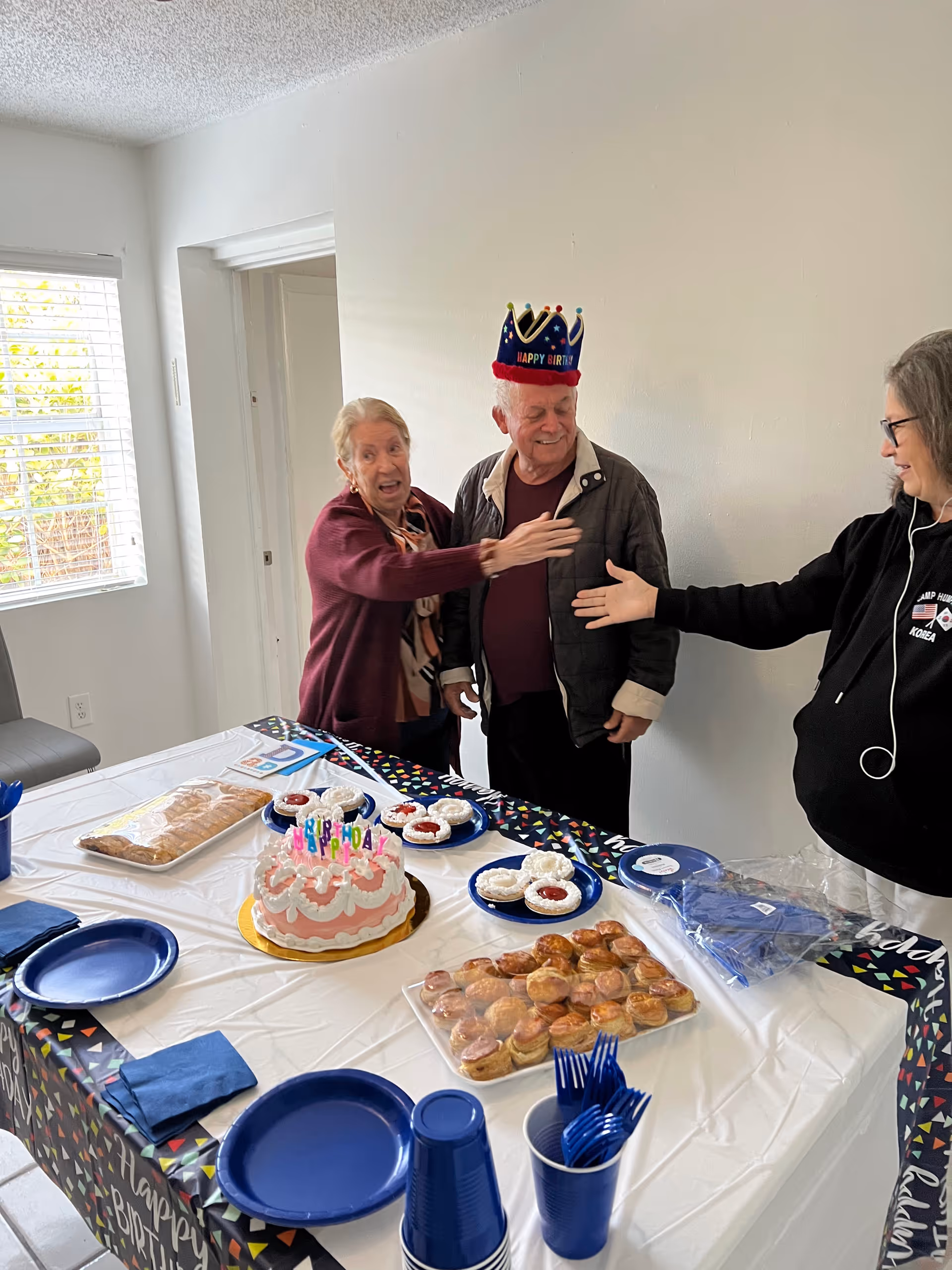 Three elderly people celebrating a birthday in a room with a table set with a birthday cake, pastries, and blue plates and cups. One man is wearing a colorful birthday crown, and two women are reaching out to greet each other. The room has white walls and a window with blinds.