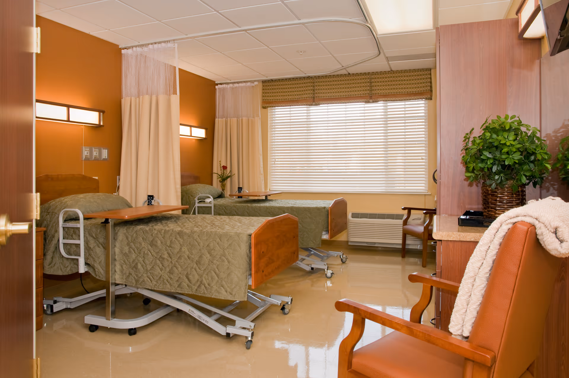A clean and well-lit room with two hospital-style beds separated by privacy curtains. Each bed has a green quilted blanket and an overbed table. There is a large window with blinds letting in natural light. The room also features wooden furniture, including a chair with a blanket draped over it and a cabinet with a potted plant on top.