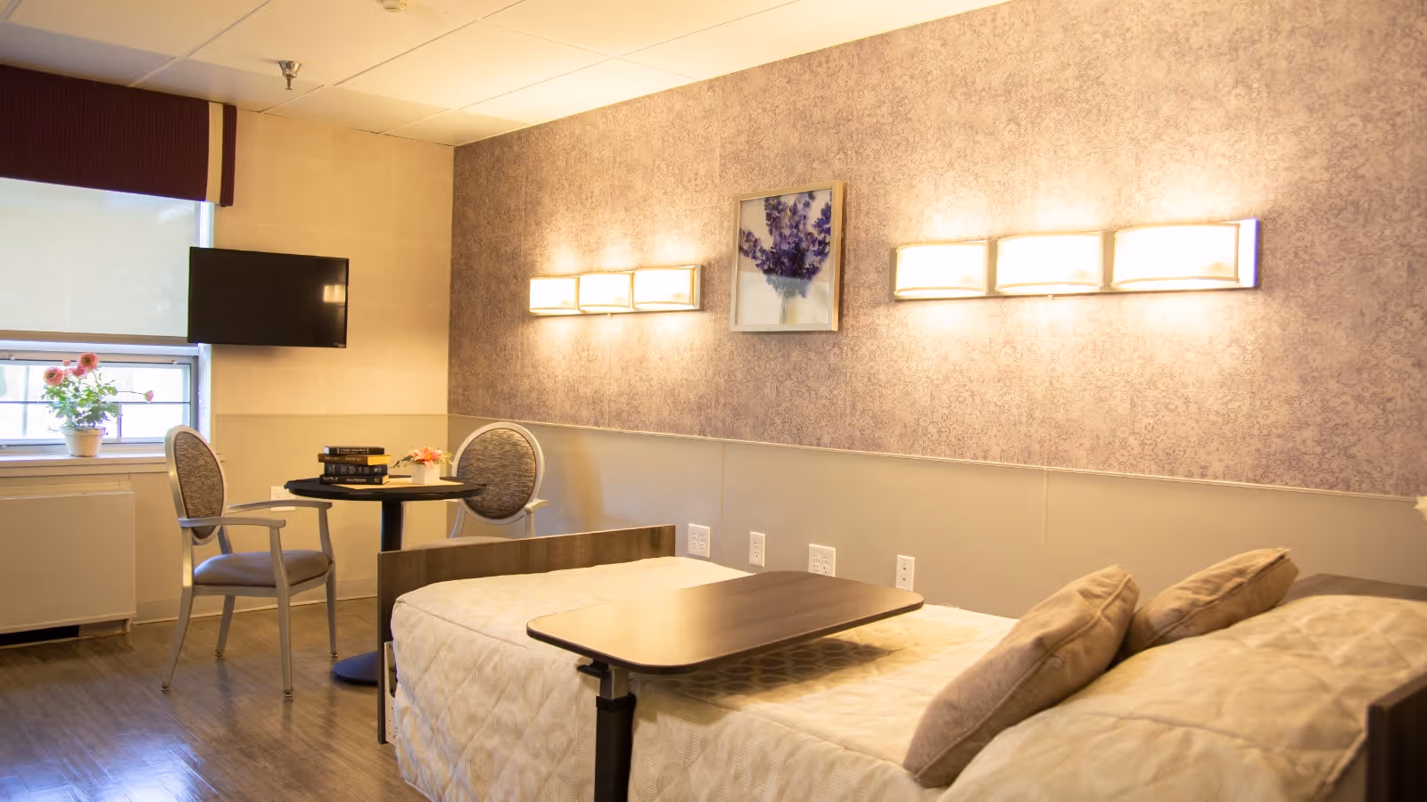 A well-lit bedroom in a senior living facility with a single bed covered in a beige quilt and two pillows. Above the bed are two modern wall lights and a framed picture of purple flowers. Next to the bed is a small round table with two chairs, a stack of books, and a small flower arrangement. A window with a roller shade and a mounted flat-screen TV are visible on the left side of the room.