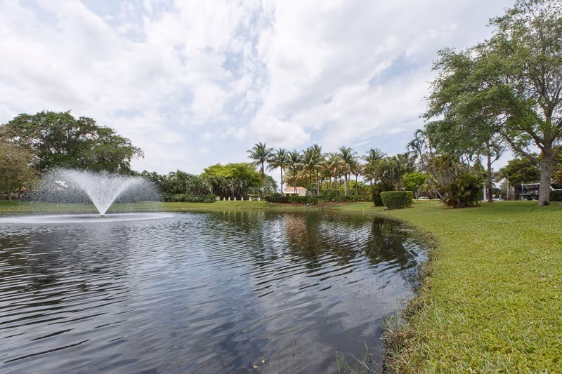 A pond with a fountain surrounded by grassy lawns, palm trees, and other trees under a cloudy sky.
